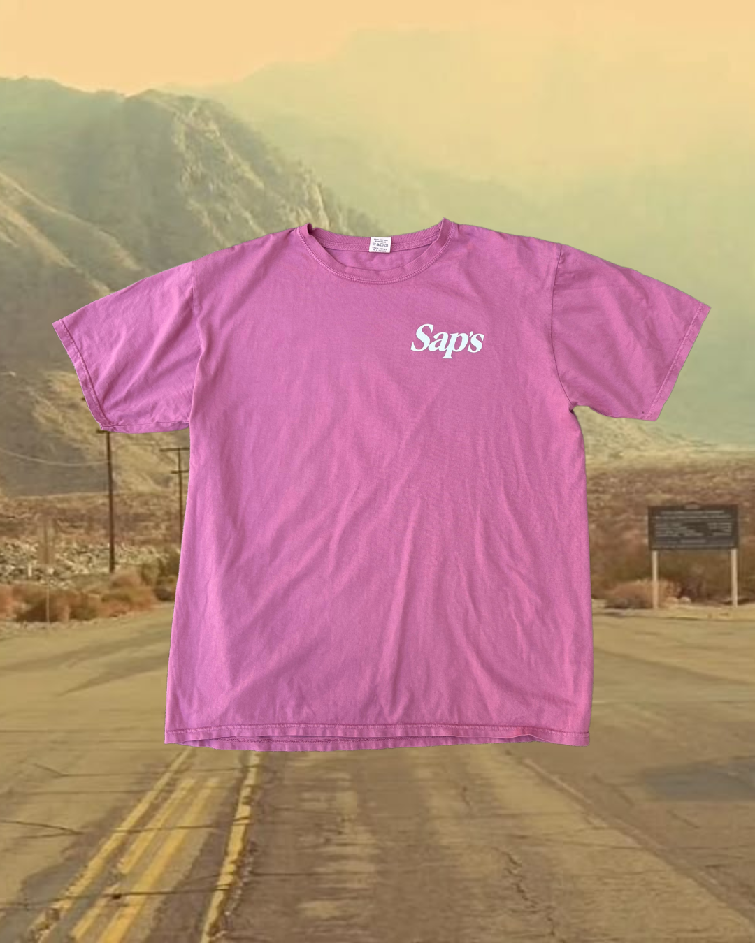 A dusty red t-shirt with the brand name 'Saps' printed on the front, displayed against a backdrop of a road leading towards mountains. #color_dusty-red