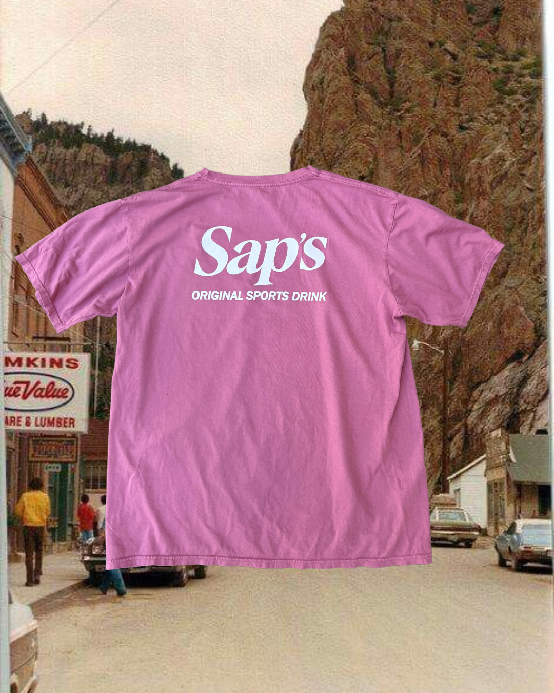 A dusty red t-shirt with 'Saps Original Sports Drink' printed on the back, displayed in a outdoor setting with a mountain and a town in the background. #color_dusty-red